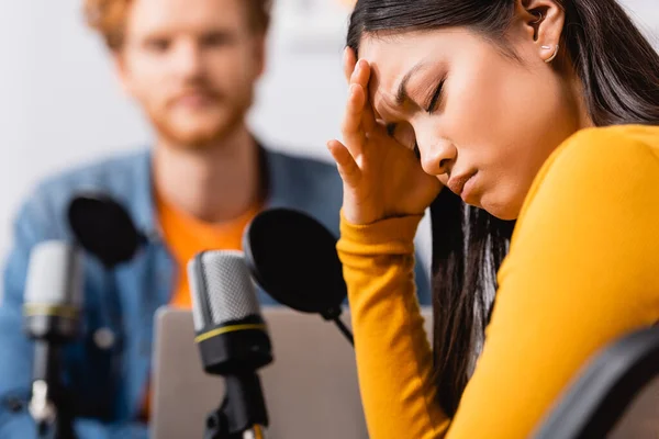 Foco seletivo de estressado asiático mulher com olhos fechados tocando cabeça durante entrevista com jovem rádio host — Fotografia de Stock