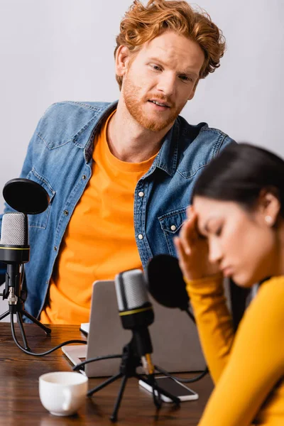 Selective focus of sad asian woman with closed eyes touching head during interview with young broadcaster — Stock Photo