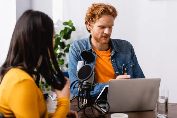 Brunette woman touching hair while sitting near microphone and redhead radio host in studio — Stock Photo