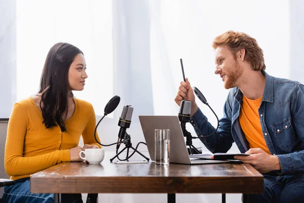 Young radio host holding pen and notebook while talking to asian woman during interview — Stock Photo