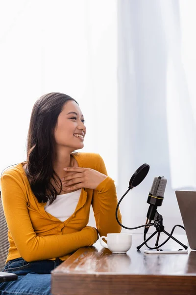 Excited asian broadcaster touching chest while sitting near microphone at workplace — Stock Photo