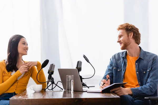 Redhead broadcaster holding notebook and pen while interviewing asian woman in studio — Stock Photo