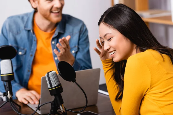 Excitada mujer asiática cubriendo la cara con la mano cerca del entrevistador en estudio de radio - foto de stock