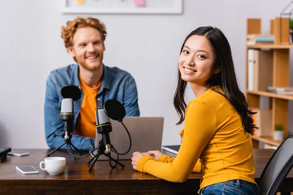 Young asian woman looking at camera near broadcaster and microphones in radio studio — Stock Photo