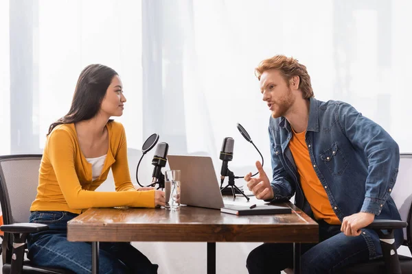 Redhead broadcaster talking to asian woman near microphones and laptop in radio studio — Stock Photo