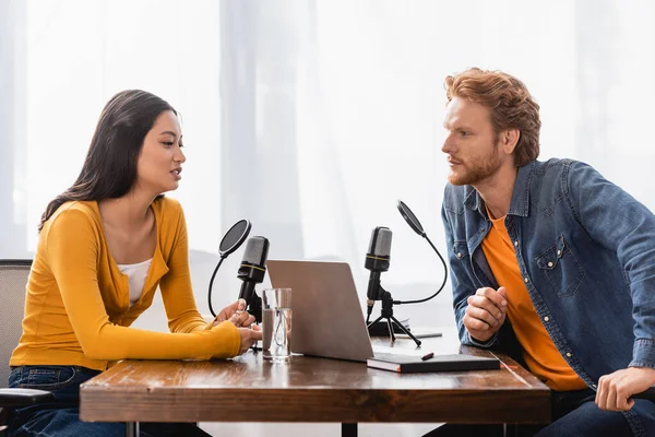 Ruiva rádio host entrevistando morena asiática mulher perto de microfones e laptop em estúdio — Fotografia de Stock