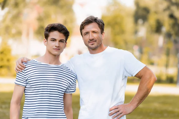 Vater steht mit Hand auf Hüfte und umarmt Teenager-Sohn draußen — Stockfoto