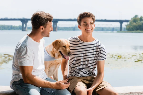 Father looking at pleased son near golden retriever and river — Stock Photo