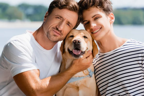 Padre e hijo mirando a la cámara y abrazando golden retriever - foto de stock