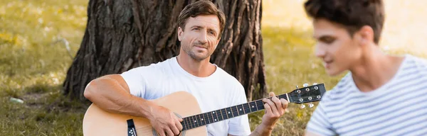 Panoramic crop of father playing acoustic guitar while looking at teenager son — Stock Photo