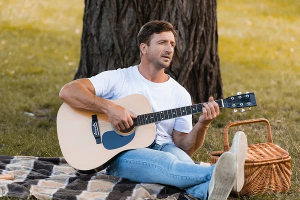 Mann sitzt auf Decke unter Baum und spielt Akustikgitarre — Stockfoto