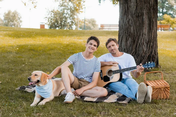 Teenager boy and father sitting on blanket near golden retriever under tree trunk — Stock Photo