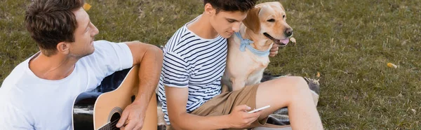 High angle view of man playing acoustic guitar near teenager boy with smartphone and golden retriever, panoramic shot — Stock Photo