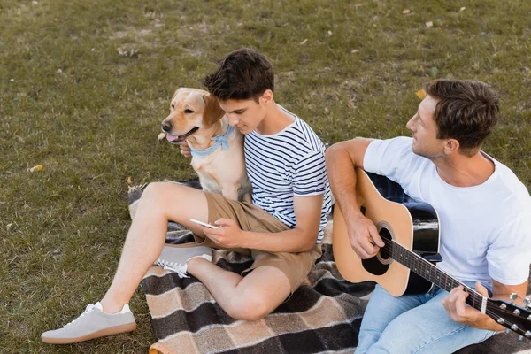 Vista de ángulo alto del hombre tocando la guitarra acústica cerca de adolescente con teléfono inteligente y golden retriever - foto de stock