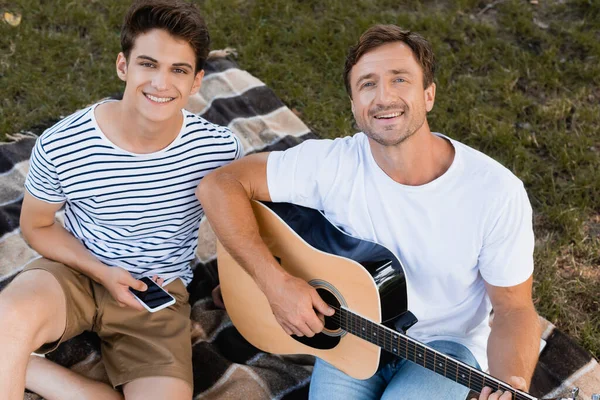 Vista ad alto angolo del padre che suona la chitarra acustica vicino al figlio adolescente con lo smartphone — Foto stock