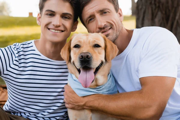 Alegre pai e adolescente menino olhando para a câmera perto golden retriever — Fotografia de Stock