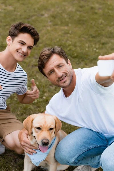 Selective focus of teenager boy pointing with finger at golden retriever while father taking selfie — Stock Photo