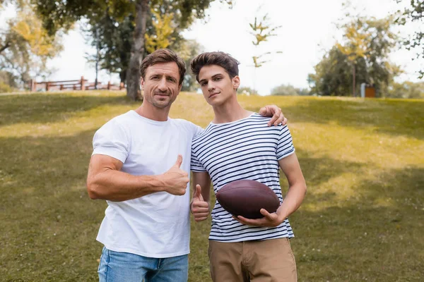 Padre e hijo adolescente con pelota de rugby mostrando pulgares hacia arriba en el parque - foto de stock