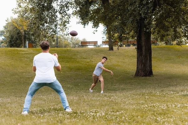 Selective focus of teenager boy playing american football with father in green park — Stock Photo