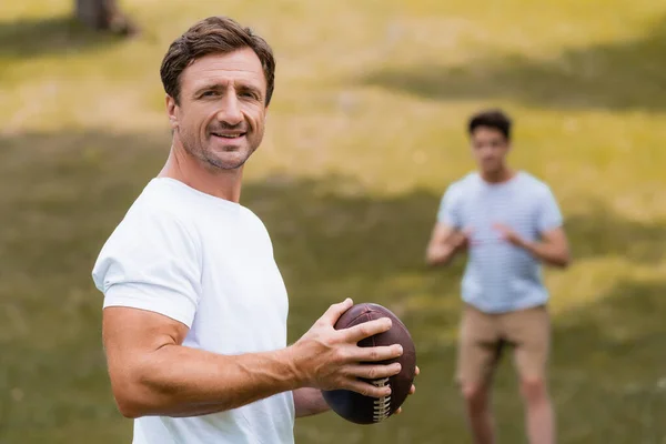 Enfoque selectivo del hombre sosteniendo la pelota de rugby y mirando a la cámara cerca de hijo adolescente en el parque verde - foto de stock