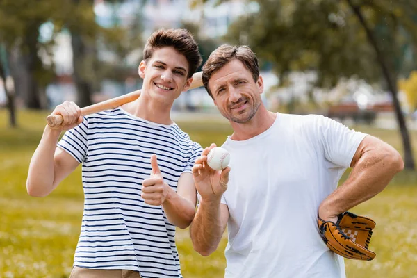 Adolescente menino com morcego softball mostrando polegar perto de pai em luva de couro segurando bola — Fotografia de Stock