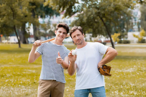 Teenager son with softball bat showing thumb up near father in leather glove holding ball — Stock Photo