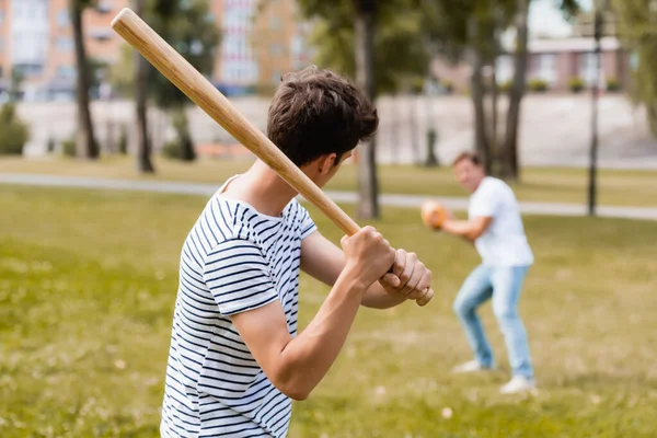 На заднем плане сын-подросток с бейсбольной битой играет в бейсбол с отцом в парке — стоковое фото