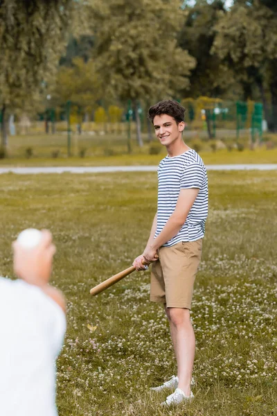 Selective focus of joyful teenager son holding softball bat near father in park — Stock Photo