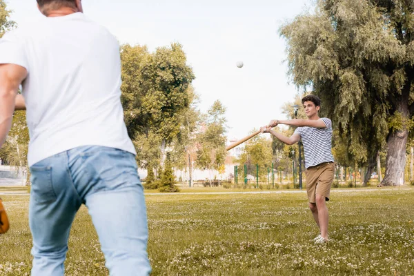 Selective focus of teenager son with softball bat looking at ball while playing baseball with father in park — Stock Photo