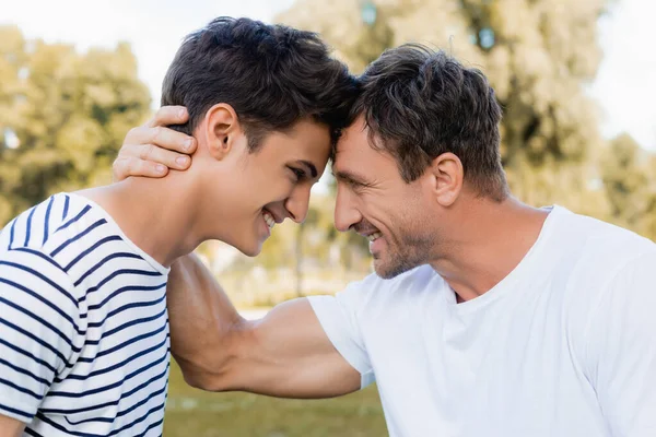 Vista lateral de alegre padre e hijo adolescente mirándose el uno al otro - foto de stock