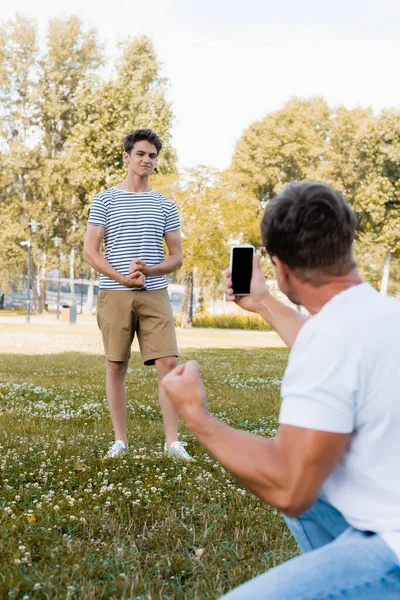 Selective focus of teenager son posing while father taking photo on smartphone in park — Stock Photo