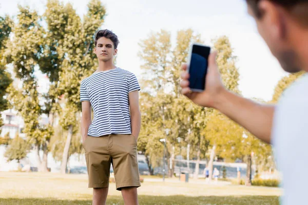 Selective focus of teenager boy standing with hands in pockets while father taking photo on smartphone in park — Stock Photo