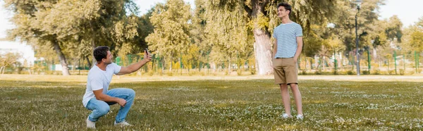 Plano panorámico del padre tomando la foto en el hijo alegre de pie con las manos en los bolsillos en el parque verde - foto de stock