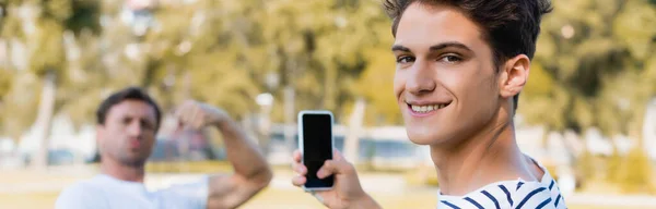 Horizontal concept of joyful teenager boy taking photo of father while holding smartphone in park — Stock Photo