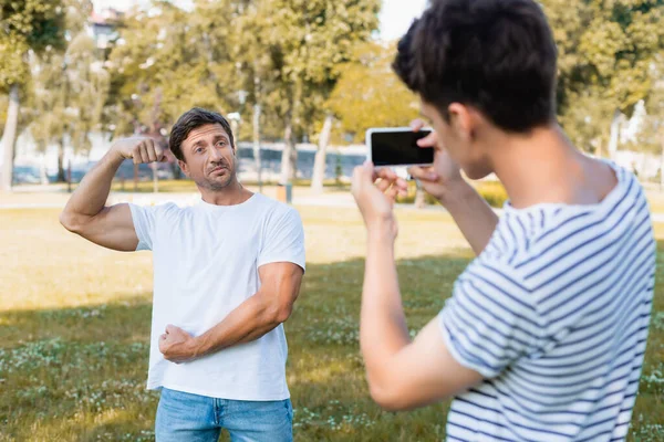 Selektiver Fokus des starken Vaters, der posiert, während Teenager-Junge Fotos macht und sein Smartphone im Park hält — Stockfoto