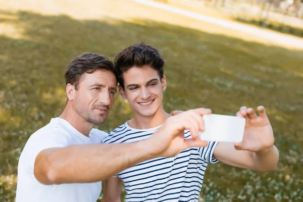 Enfoque selectivo de padre e hijo adolescente tomando selfie en parque - foto de stock