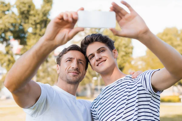 Foco seletivo de pai satisfeito e filho adolescente tomando selfie no parque — Fotografia de Stock