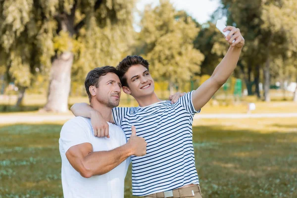 Padre mostrando el pulgar hacia arriba mientras adolescente hijo tomando selfie en parque - foto de stock