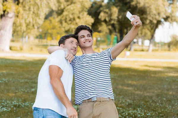 Teenager son hugging father while taking selfie in green park — Stock Photo