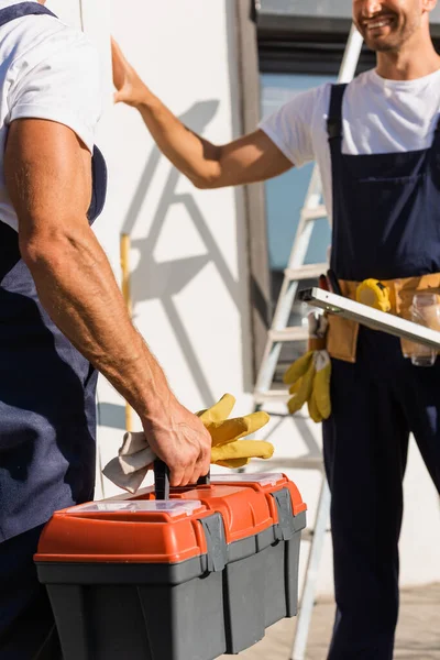Vista recortada del constructor sosteniendo guantes y caja de herramientas cerca de colega al aire libre — Stock Photo