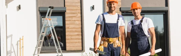 Horizontale Ernte von Bauarbeitern in einheitlichen Haltewerkzeugen und Bauplänen in der Nähe von Gebäuden — Stockfoto