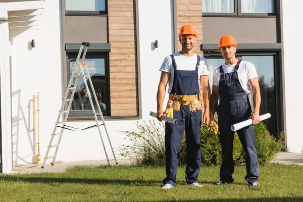 Bauarbeiter in Arbeitskleidung halten Wasserwaage und Bauplan auf Rasen neben Gebäude — Stockfoto