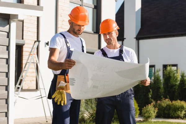Arbeiter in Overalls und Helmen betrachten den Bauplan in der Nähe des Gebäudes — Stockfoto