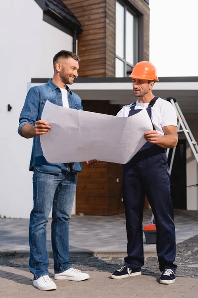 Bauunternehmer in Arbeitskleidung hält Bauplan mit Kunden in der Nähe des Gebäudes — Stockfoto