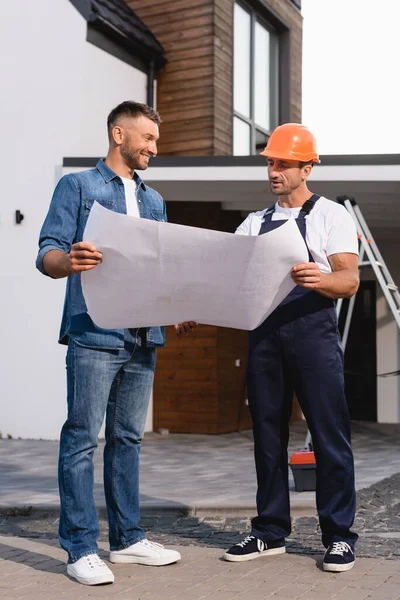 Costruttore in abbigliamento da lavoro che tiene il progetto con il cliente vicino all'edificio — Foto stock