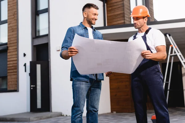 Workman in helmet holding blueprint near man and building outdoors — Stock Photo