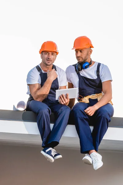 Constructores que utilizan tableta digital mientras están sentados en el techo del edificio aislado en blanco - foto de stock