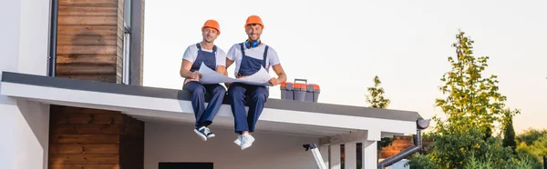 Imagen panorámica de constructores en uniforme sosteniendo el plano cerca de la caja de herramientas en el techo del edificio - foto de stock