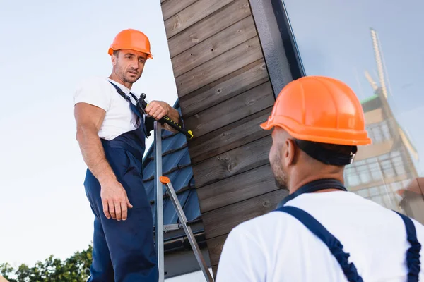Selective focus of builder holding hammer while standing on ladder near colleague and facade of building — Stock Photo