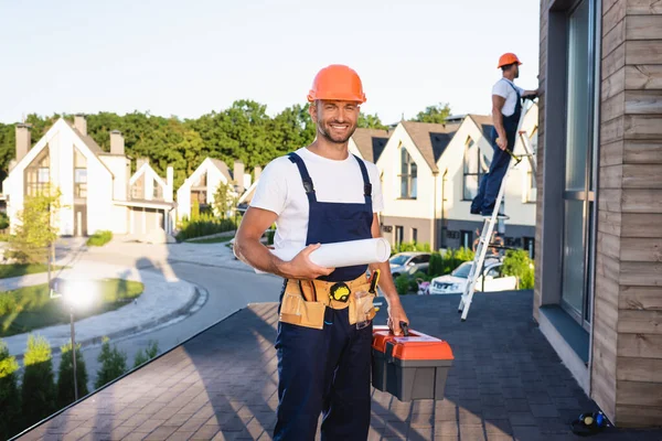 Focus selettivo del costruttore con cassetta degli attrezzi e cianografia guardando la fotocamera mentre il collega lavora sul tetto — Foto stock